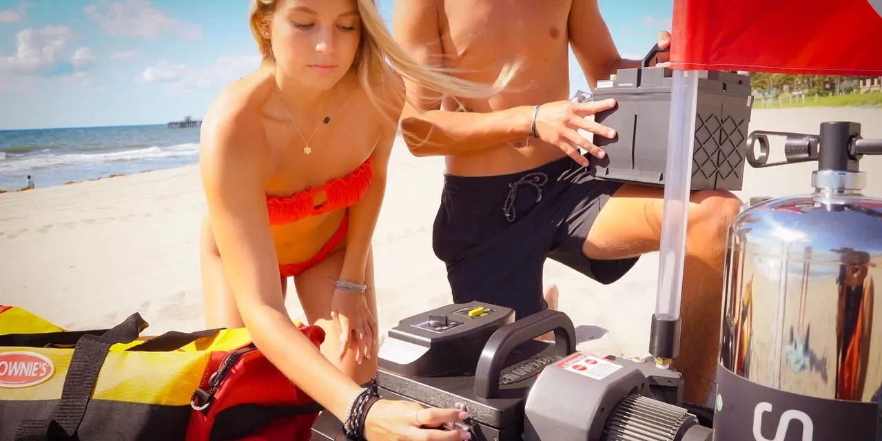 821673a5-e967-4a55-8e0e-513422e5254c.webp A woman and man prepare a Brownie’s Third Lung tankless dive system on a sandy beach, assembling the compressor and battery next to a red-and-yellow dive gear bag and a dive flag, with the ocean and pier visible in the background.