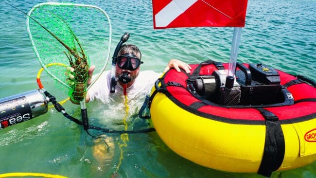 Diver using a Brownie’s Sea LiON floating hookah diving system to catch a lobster near the surface, with red and yellow float, dive flag, and battery-powered compressor.
